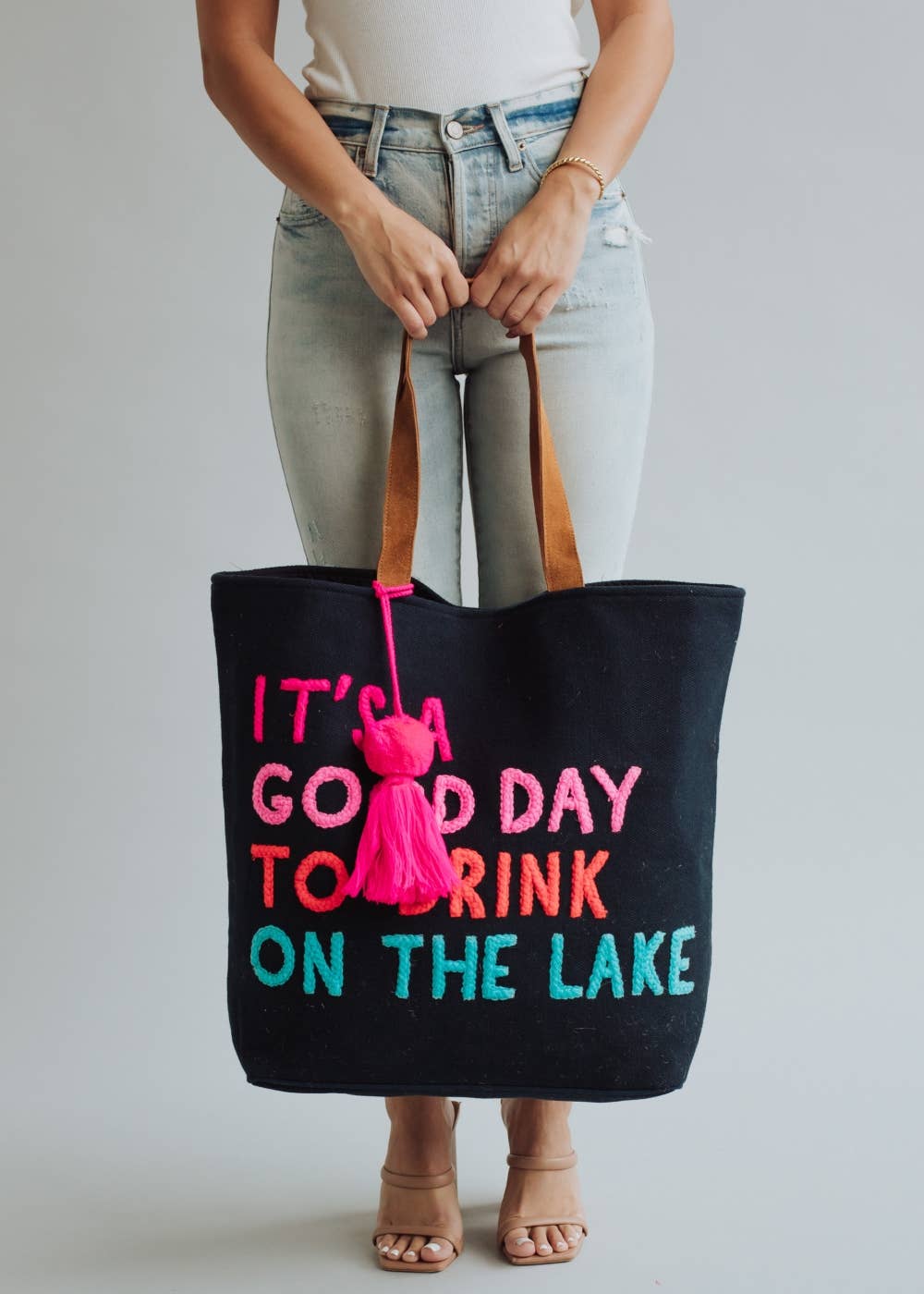 Black tote bag with colorful text 'It's a good day to drink on the lake' held by a person.